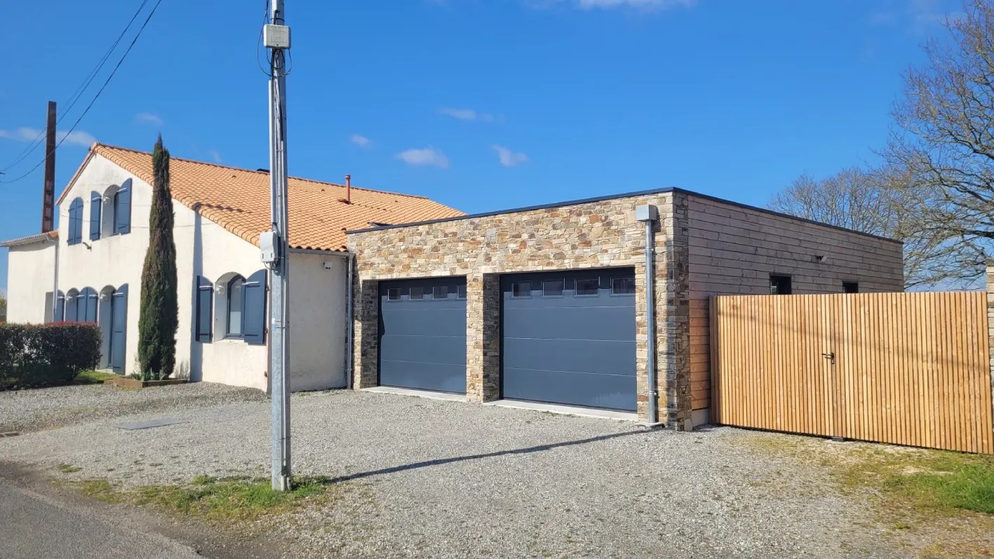 Extension de la maison : préau fermé côté rue par des portails sectionnels en métal laqué, mur en ossature bois et mur en pierres apparentes